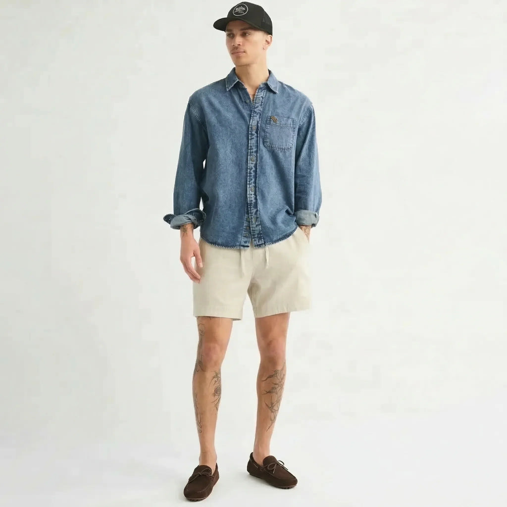 Man wearing denim shirt, beige shorts, brown loafers, and a black cap on white background