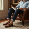 Man in casual jeans and shirt wearing brown leather loafers, sitting in a leather chair and reading a book.