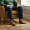 Men's cognac leather slip-on shoes with jeans, seated on a tan armchair indoors