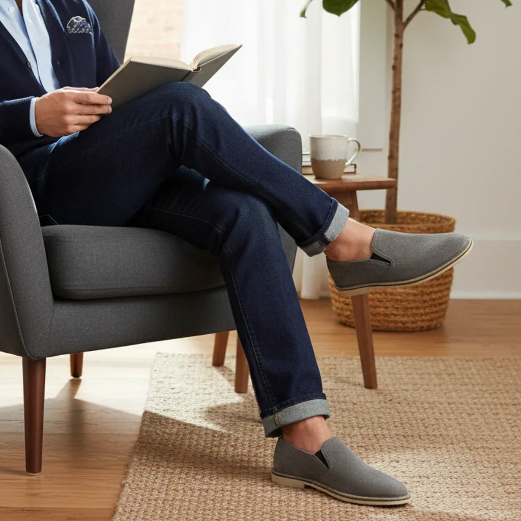 Man wearing gray slip-on shoes, dark jeans, sitting in a chair reading in a cozy living room